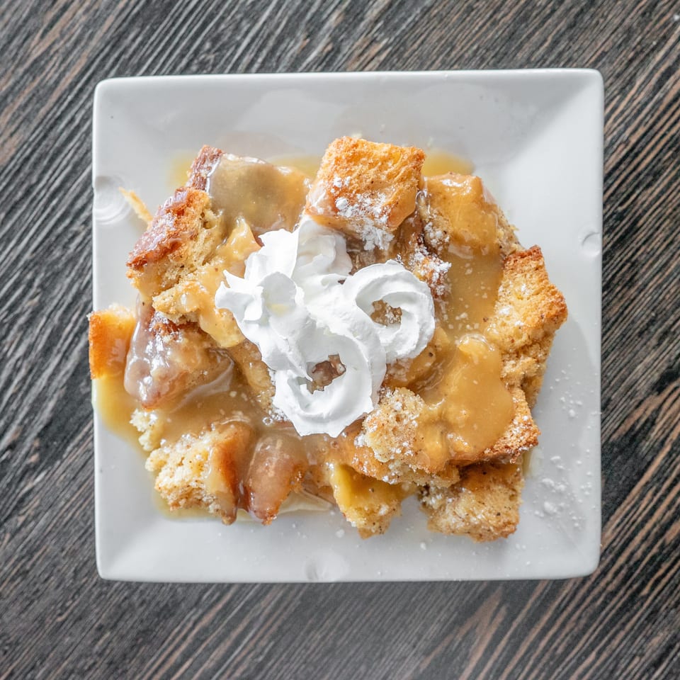 Golden bread pudding topped with caramel sauce, powdered sugar, and whipped cream on a square white plate.
