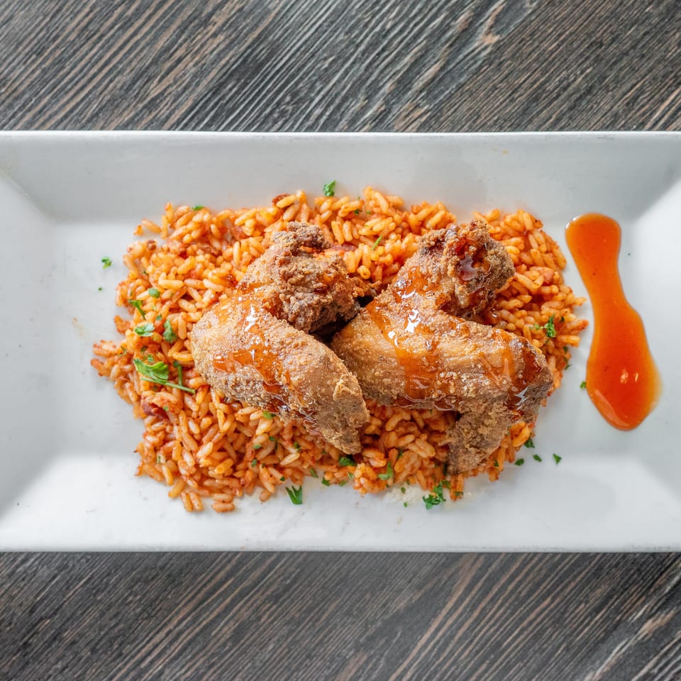 Crispy fried chicken wings glazed with sauce over seasoned red rice, garnished with herbs and served with extra sauce.