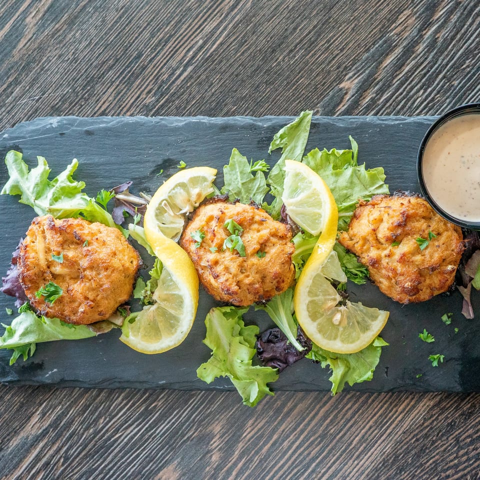 Three golden-brown fritters with lemon wedges and leafy greens on a slate platter, accompanied by creamy dipping sauce.