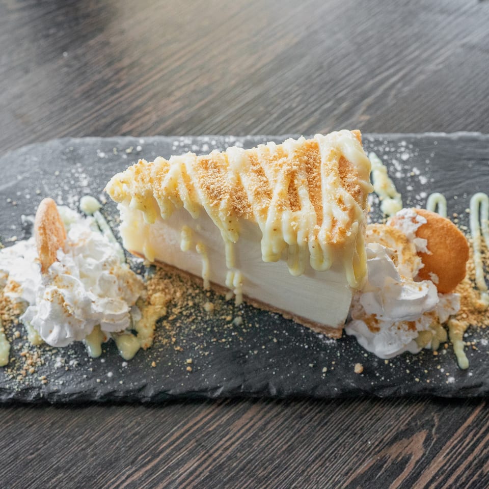 Slice of creamy layered cake topped with caramel drizzle and crumbs, surrounded by whipped cream and pastry pieces on a black slate plate.