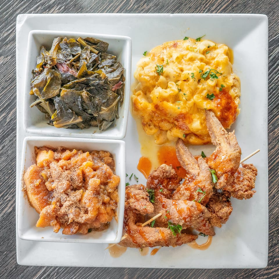 Overhead view of a platter with macaroni and cheese, leafy greens, sweet potatoes, and sauced fried chicken skewers.
