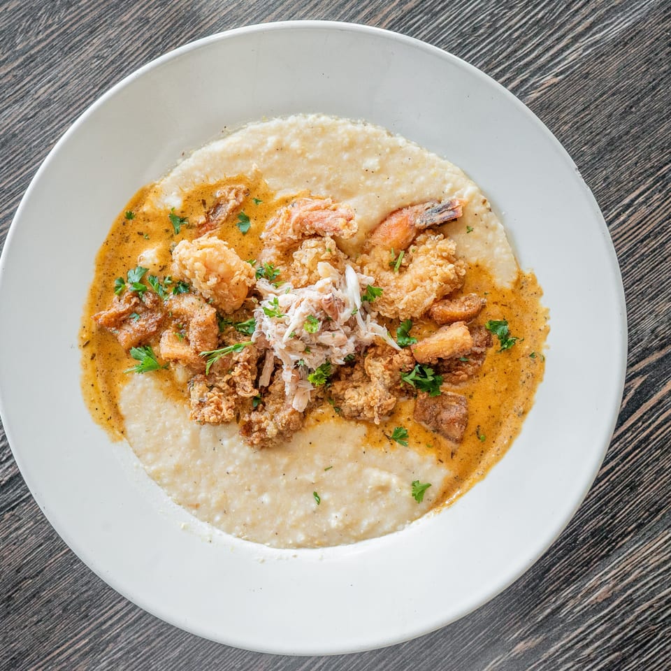 Creamy grits topped with fried shrimp, crab meat, savory sauce, and parsley on a white plate.