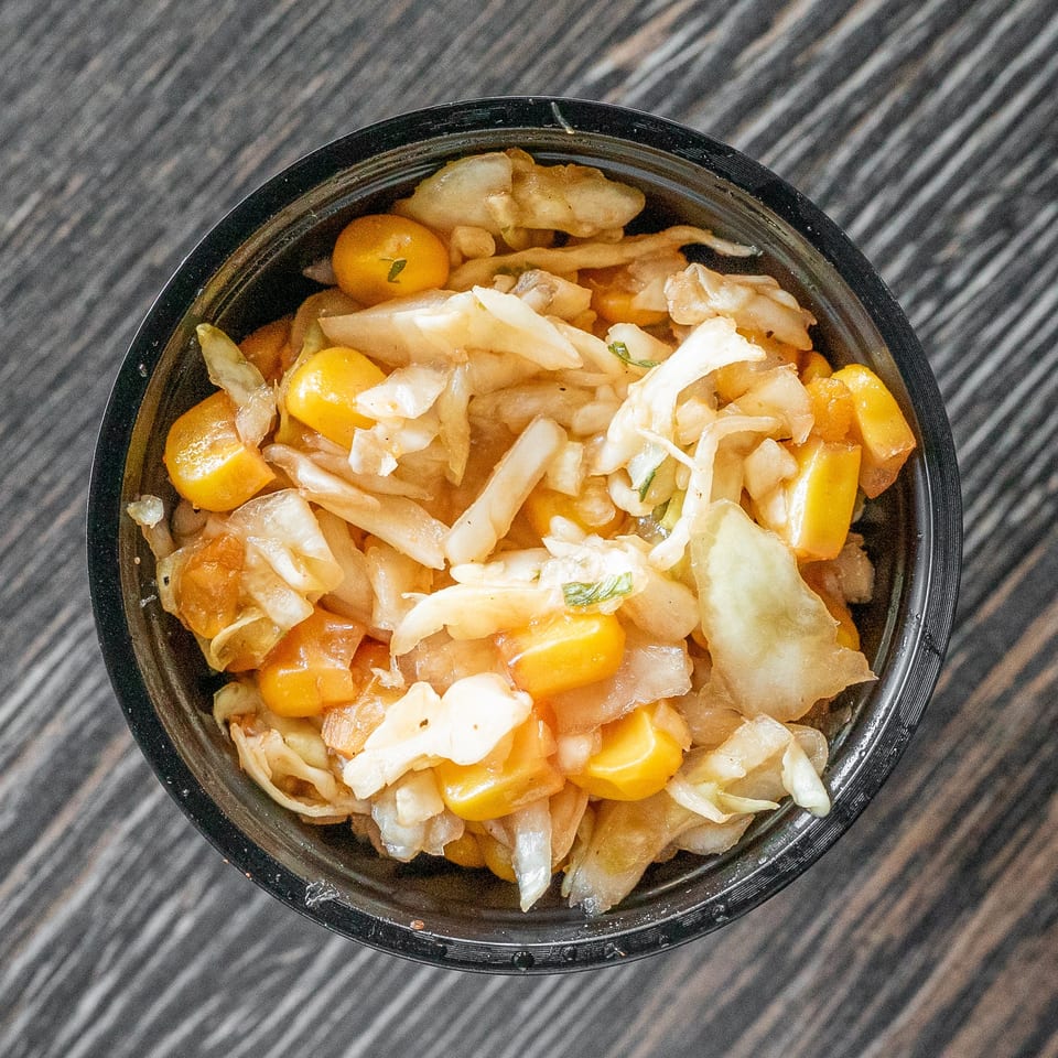 Overhead view of a black bowl filled with chopped cabbage, yellow corn, tomatoes, and other vegetables on a dark wood table.