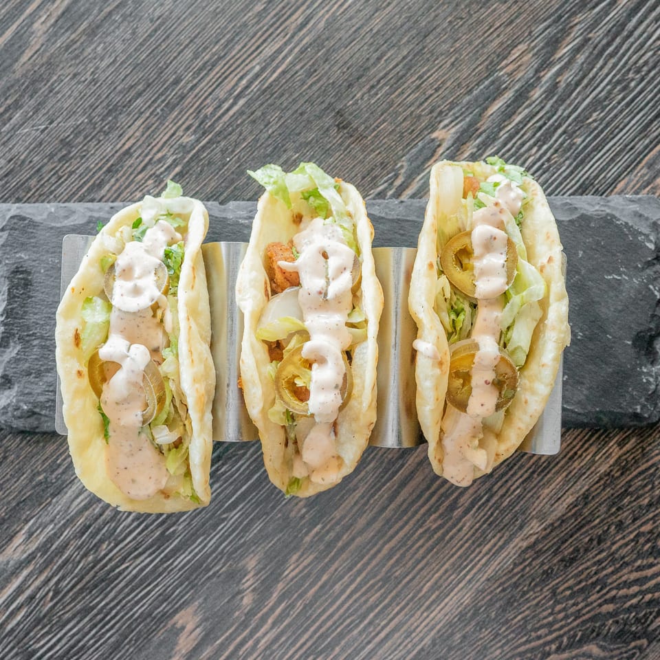 Three soft tacos filled with lettuce, jalapeños, and creamy sauce, arranged in a metal holder on a slate serving board.