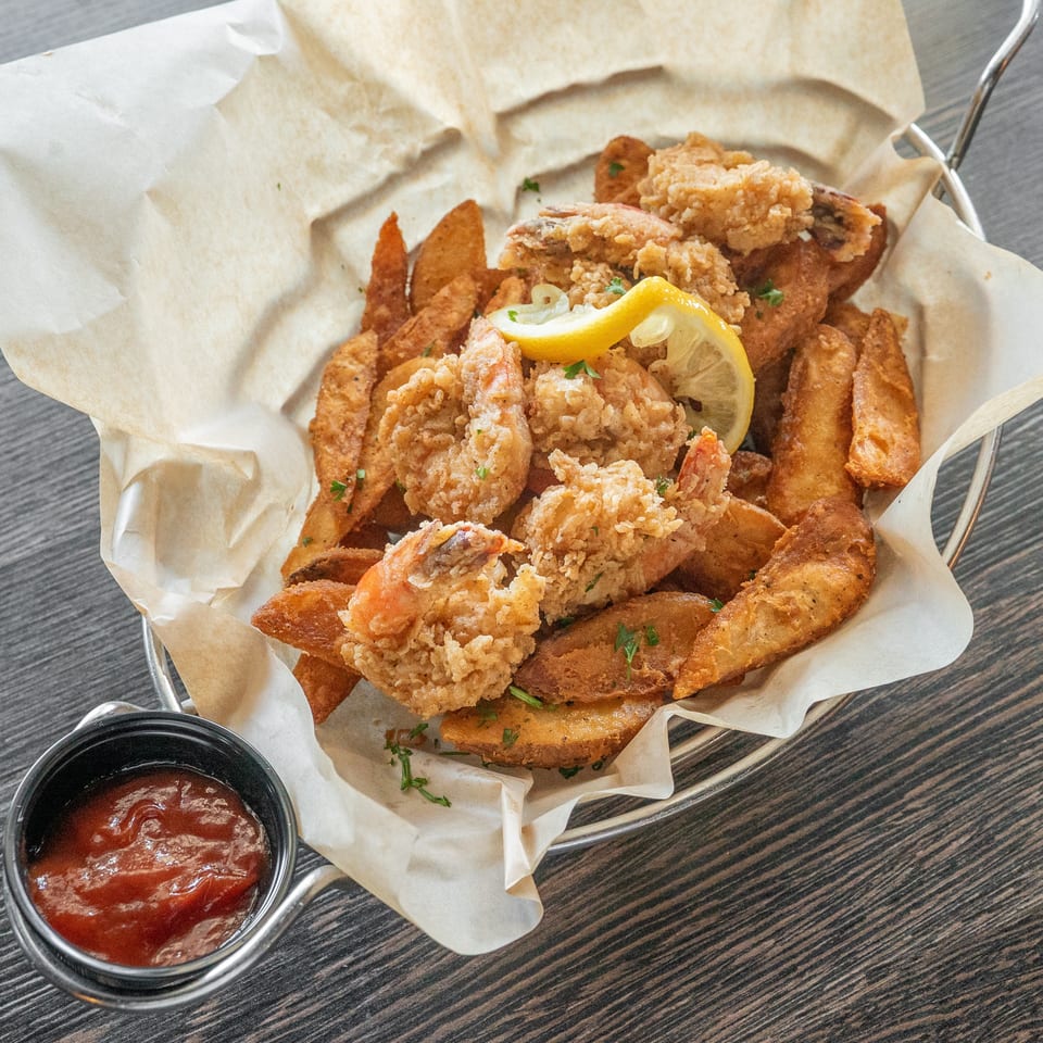 Basket of crispy fried shrimp and potato wedges garnished with parsley and lemon, served with dipping sauce.