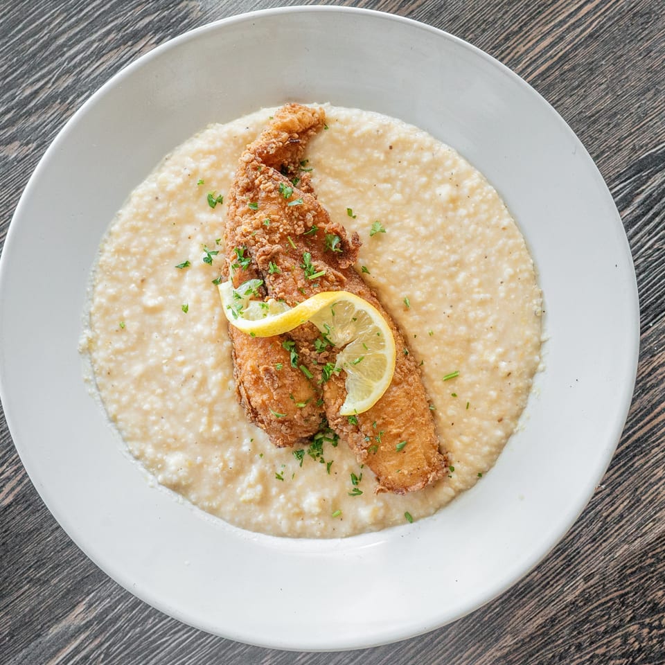 Crispy fried fish fillets with lemon and parsley served over creamy grits on a white plate.