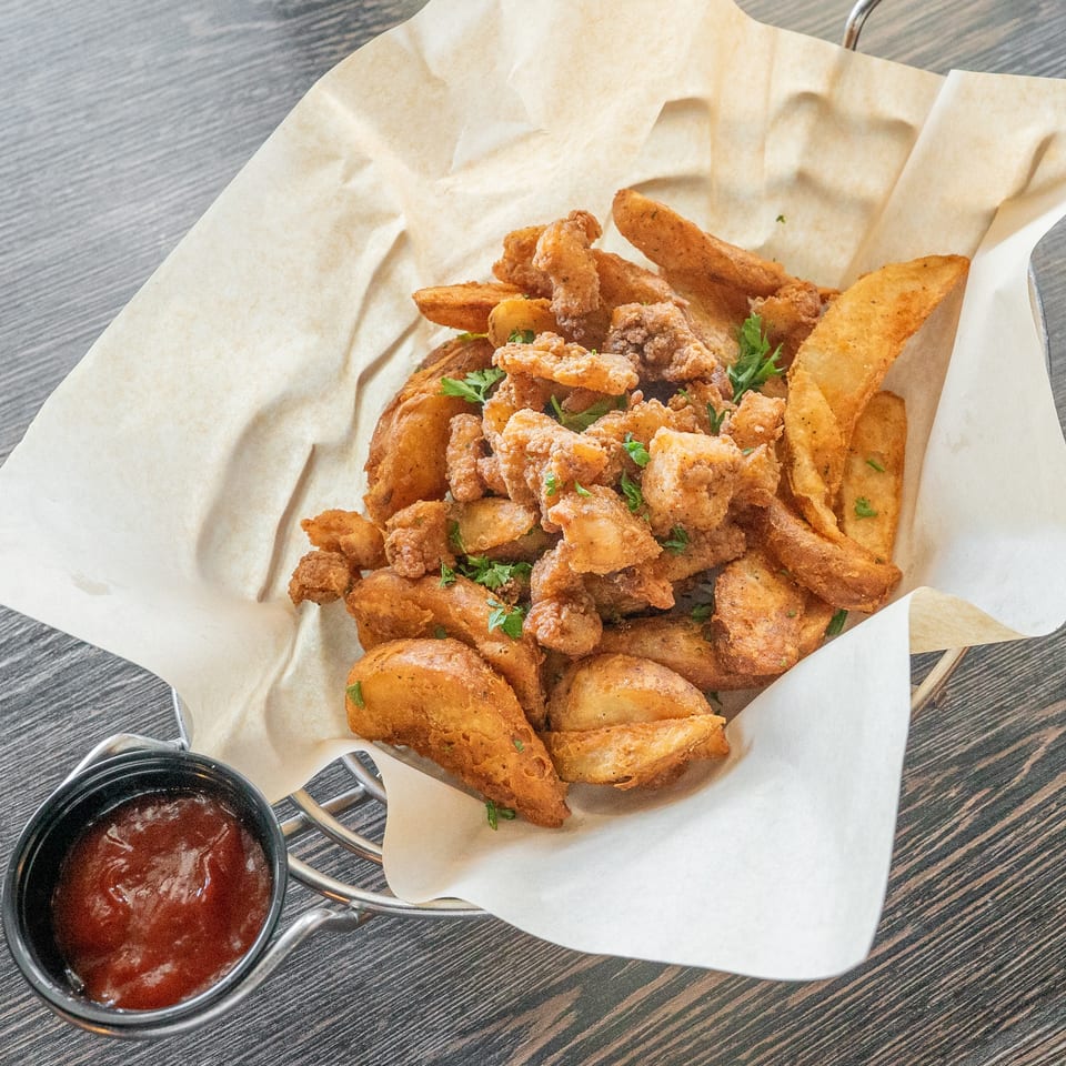 Basket of fried chicken pieces and potato wedges garnished with parsley, served with dipping sauce on a paper-lined tray.