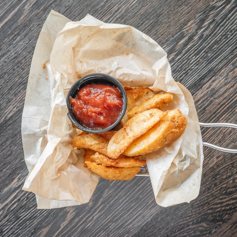 Golden fried potato wedges served with a cup of red dipping sauce on parchment paper over a dark wooden table.