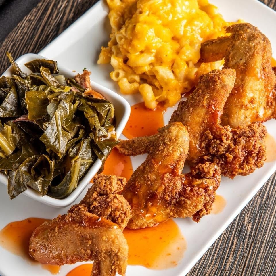 Plate of fried chicken wings with macaroni and cheese, collard greens, and orange sauce on a wooden table.