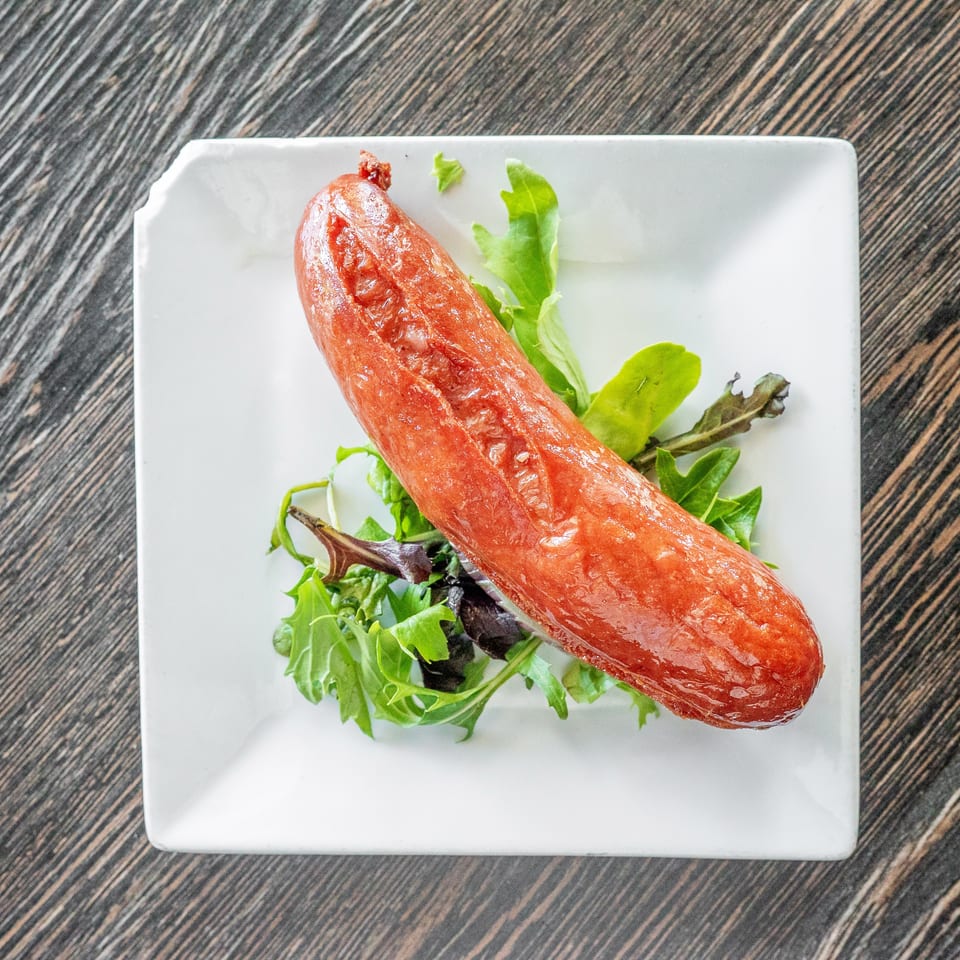Glossy grilled sausage served diagonally over mixed greens on a white square plate.