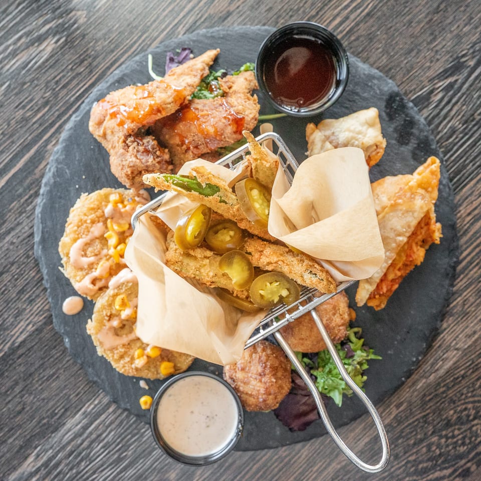 Assorted crispy fried appetizers with jalapeños, sauces, and greens arranged on a black slate platter.