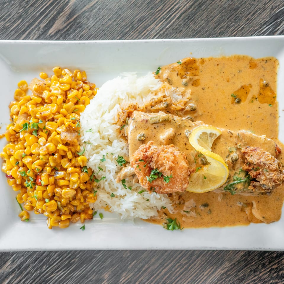 Breaded fish with lemon and creamy caper sauce, white rice, and seasoned corn served on a rectangular white plate.