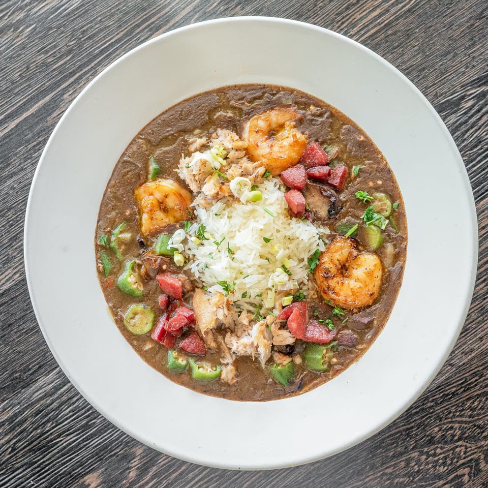 White rice topped with shrimp, crab, sausage, okra, and vegetables in a dark savory sauce on a white plate.