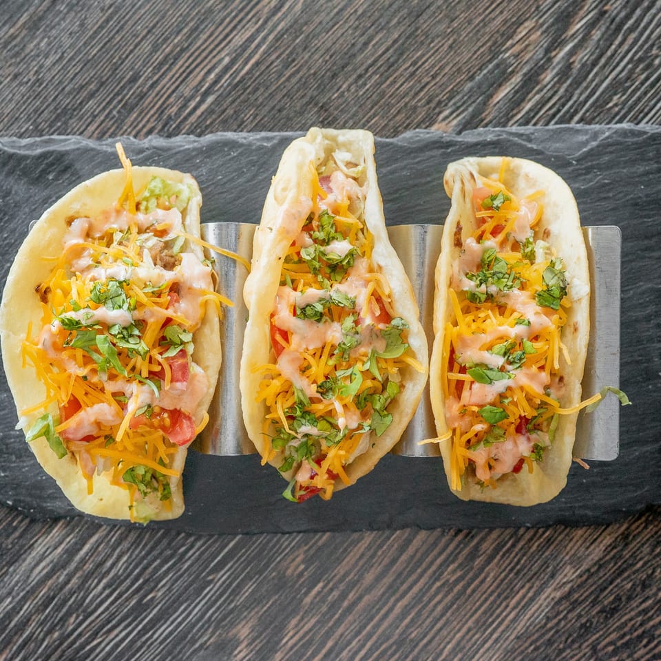 Three soft tacos topped with shredded cheddar cheese, chopped cilantro, diced tomatoes, lettuce, and creamy sauce on a slate platter.
